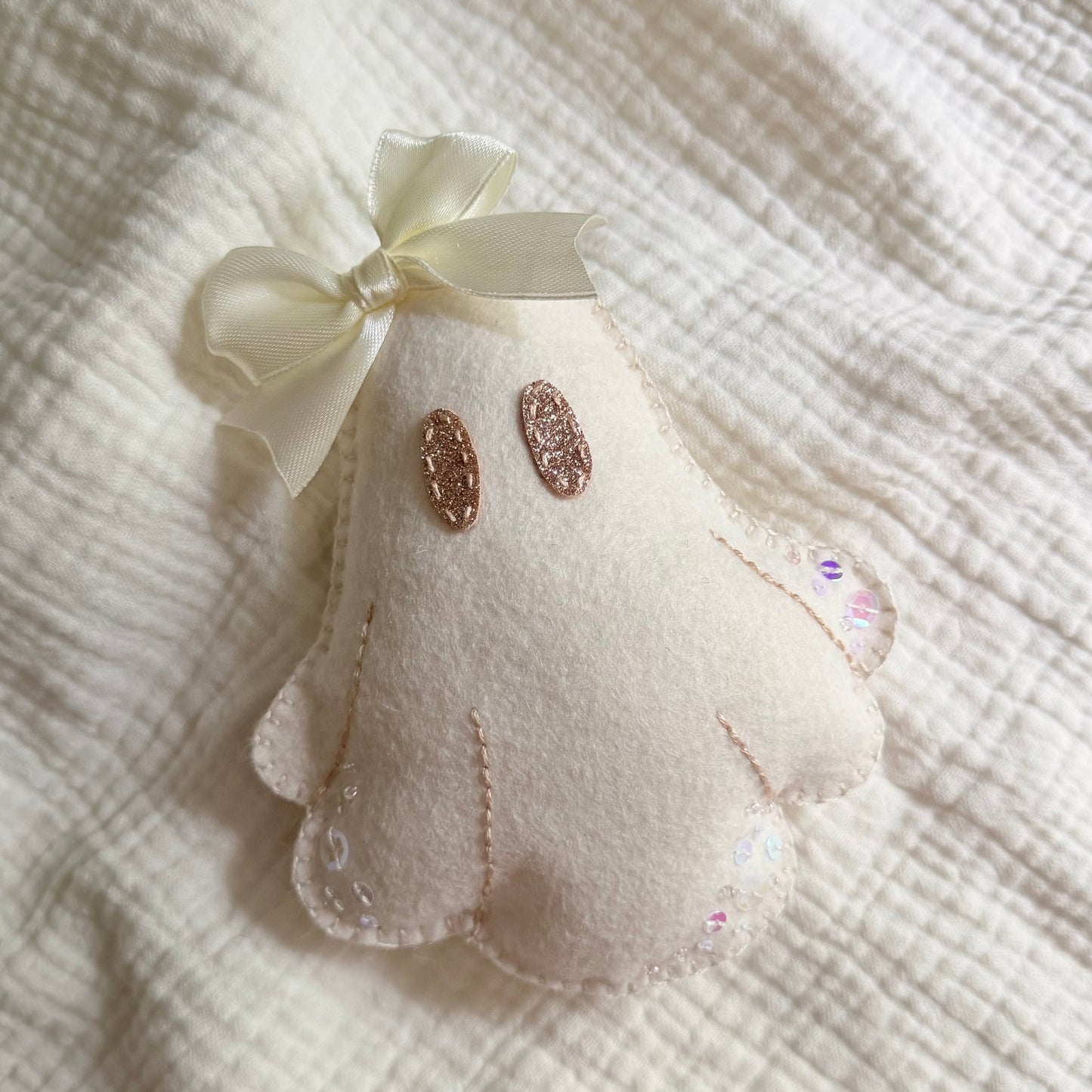 A small cream-colored felt ghost ornament with embroidered details, a cream satin bow, and glittery rose-gold eyes, resting on a textured cream gauze fabric background.