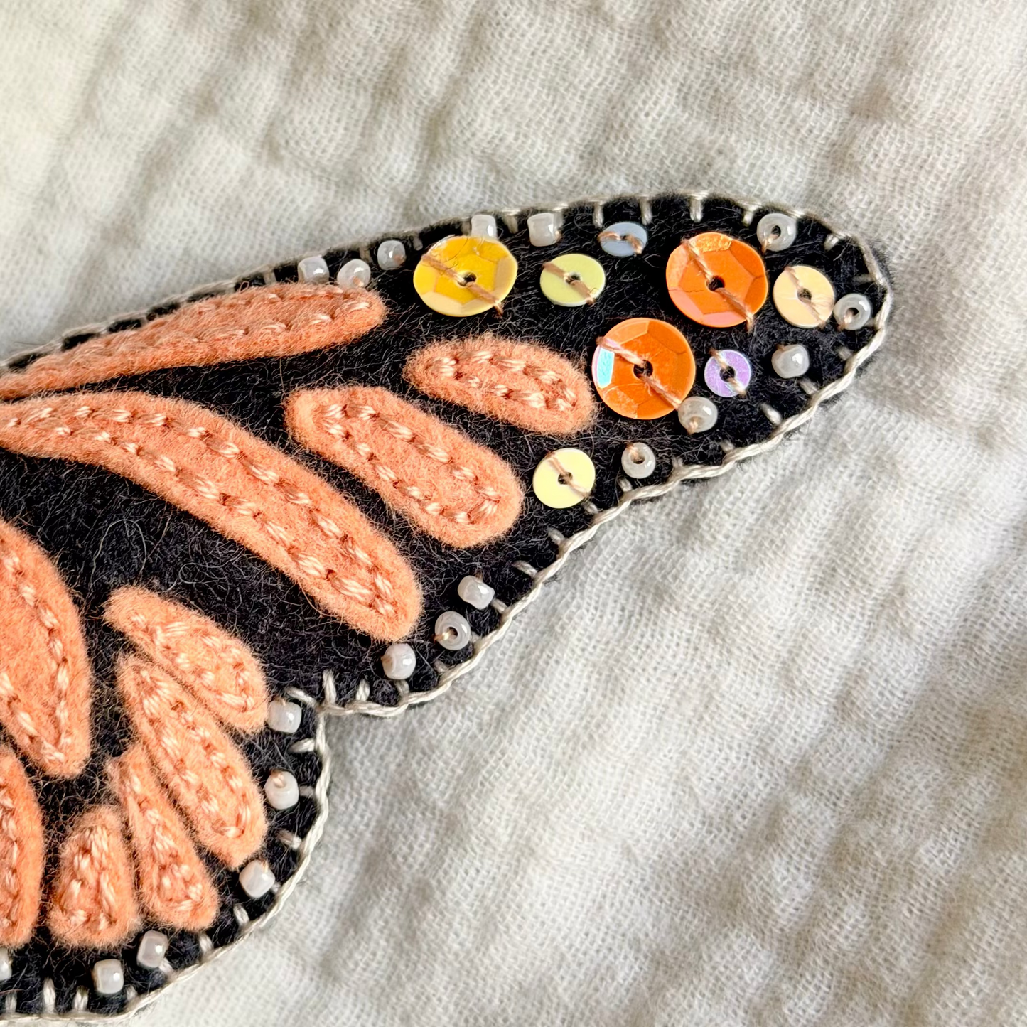 A close-up of a monarch butterfly ornament with orange and black embroidered wings, decorated with small colorful sequins, resting on a textured cream fabric background.