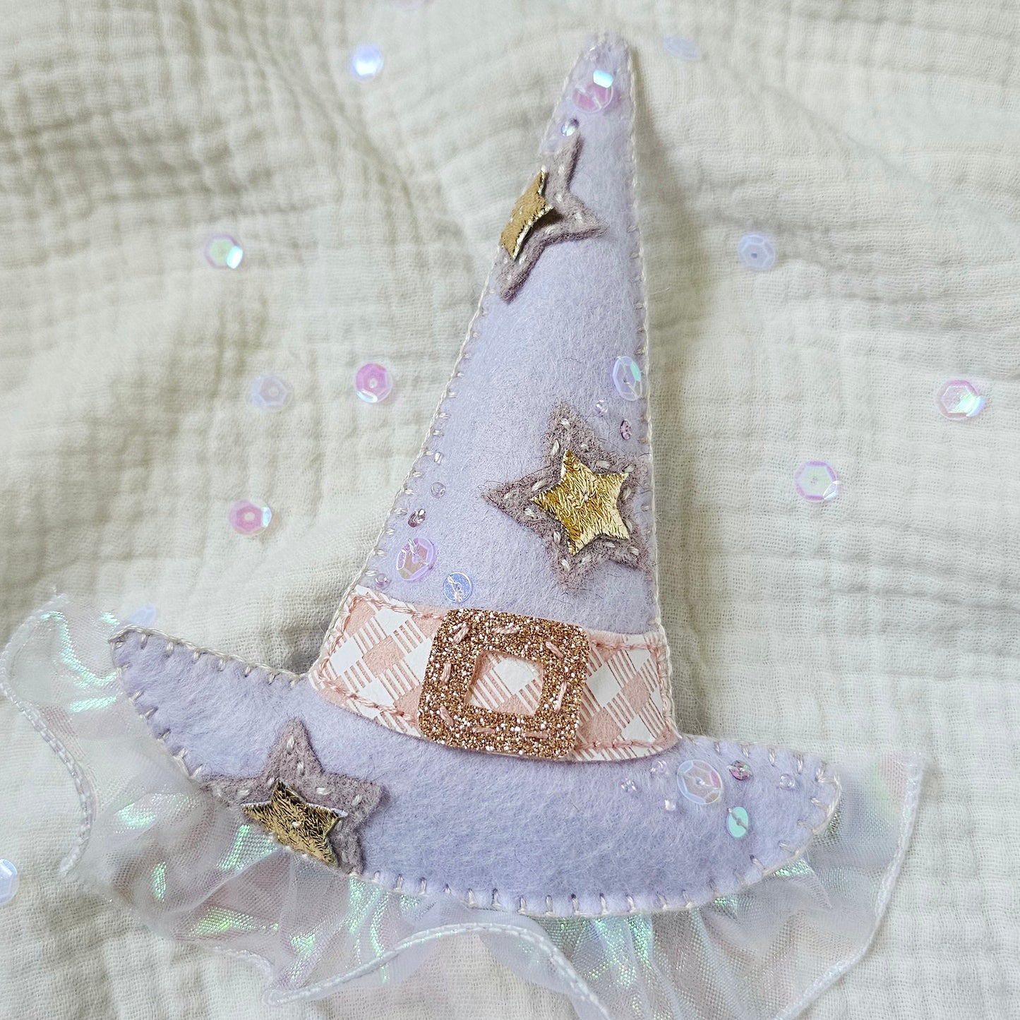 A small lavender fabric witch hat applique with embroidered gold stars and a glittery gold band, placed on a textured cream gauze fabric background with scattered iridescent sequins.