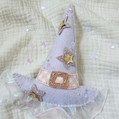 A small lavender fabric witch hat applique with embroidered gold stars and a glittery gold band, placed on a textured cream gauze fabric background with scattered iridescent sequins.