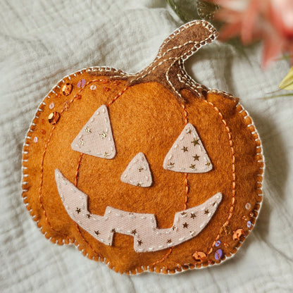 A handmade orange felt jack-o'-lantern ornament with a stitched stem and carved face, decorated with small sequins, resting on a textured white fabric with blurred flowers in the background.