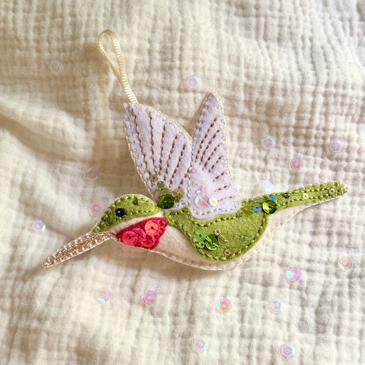 A handmade felt hummingbird ornament with green body, white wings, pink throat, and sequined details, resting on a textured cream gauze fabric background.