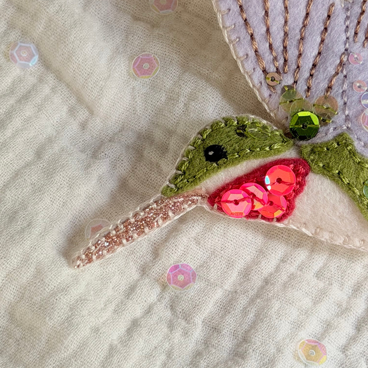 A close-up of a handmade felt hummingbird applique on a textured cream fabric background with scattered sequins. The applique features a lavender wing, green body, pink glitter beak, and sequin details.