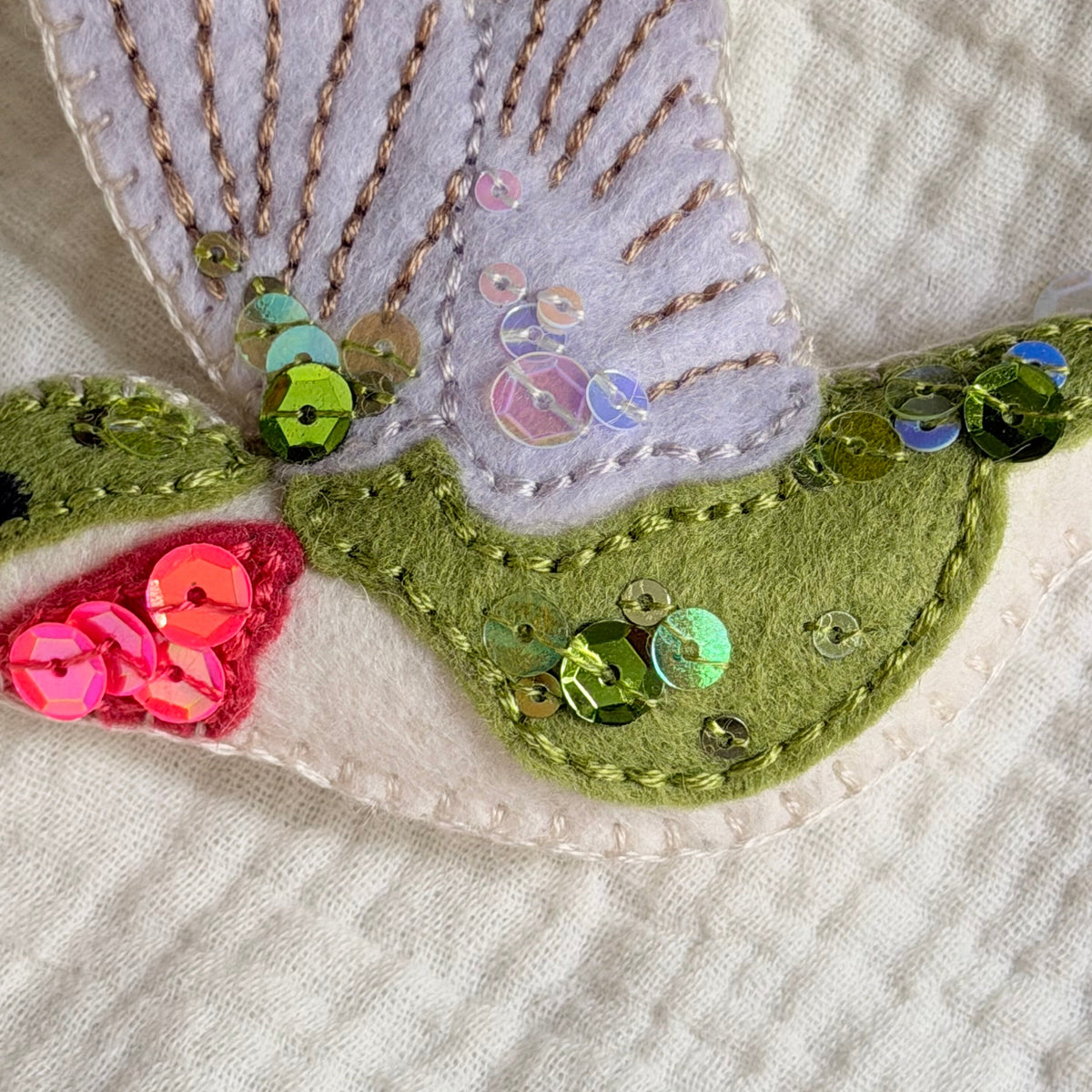Close-up of a handmade felt applique featuring a butterfly wing in pale lavender with gold thread embroidery, an olive green body with iridescent sequins, and a pink throat detail on a cream quilted fabric background.