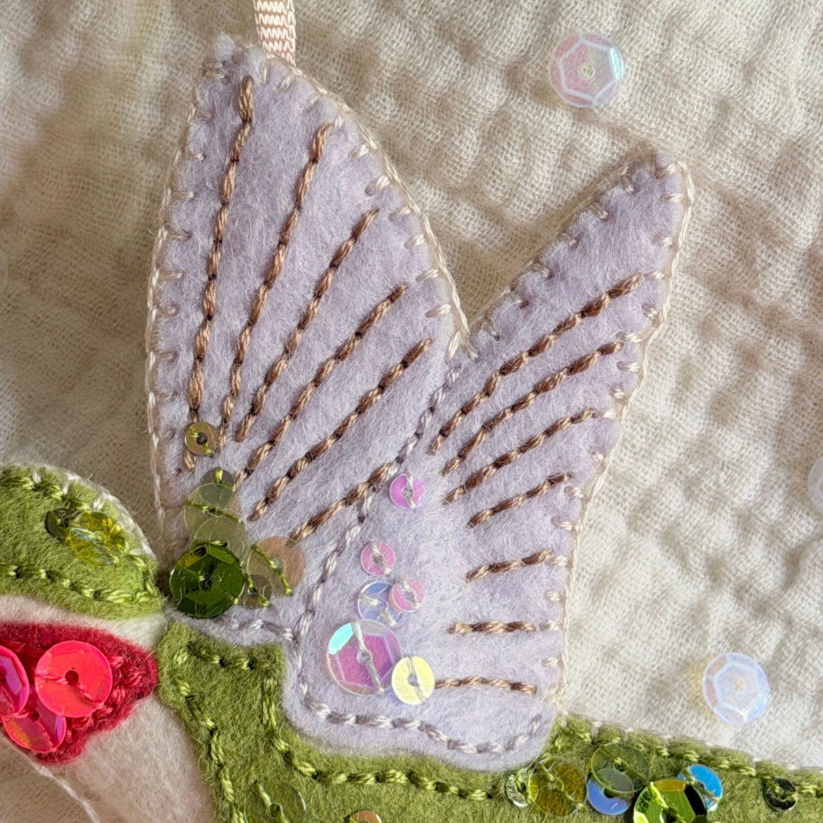 Close-up of a handmade felt hummingbird ornament featuring a pale lavender wing with gold embroidery, green felt body details, iridescent sequins, and a cream ribbon loop at the top.