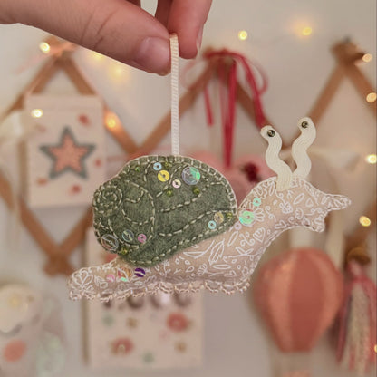 A handmade snail ornament being held by a hand, featuring a green embroidered shell with sequins and a white lace body, set against a softly blurred festive background with warm lights.