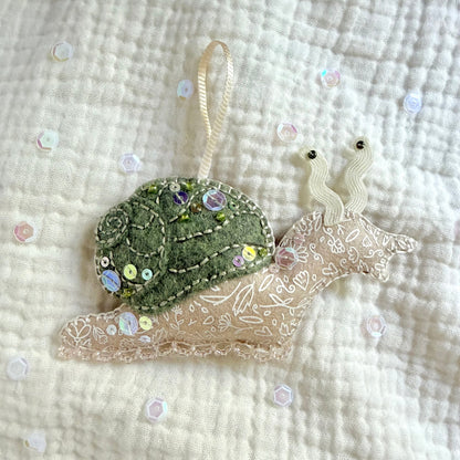 A handmade snail ornament or decoration featuring a green sequined shell and a beige embroidered body, resting on a textured cream muslin fabric with scattered sequins.
