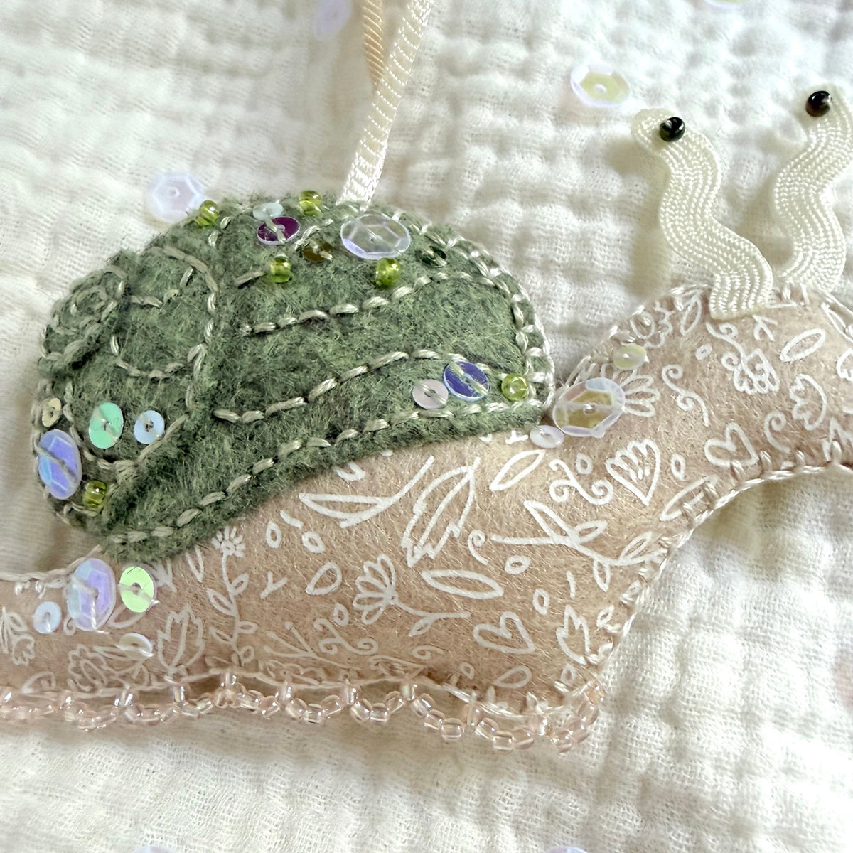 A close-up of a handmade fabric ornament shaped like a snail, featuring a textured olive green felt shell with embroidered leaf patterns and iridescent sequins, and a beige fabric body with white floral embroidery and lace trim, resting on a quilted cream fabric background.