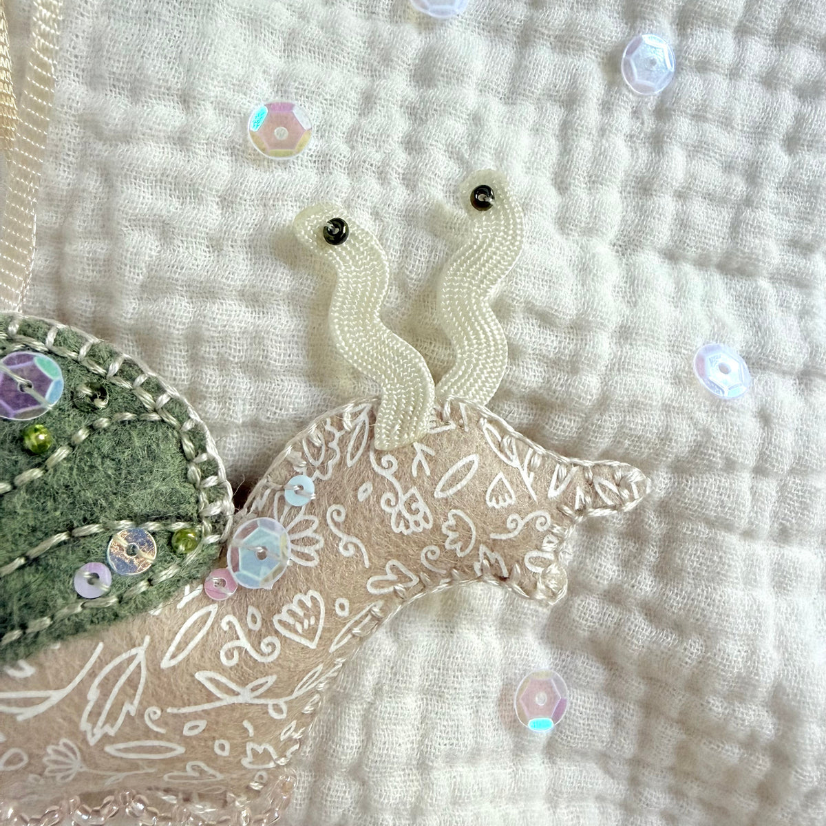 A close-up of a handmade fabric ornament shaped like a snail, featuring a beige embroidered body, a green sequined shell, and cream ribbon antennae, displayed on a textured white fabric background.