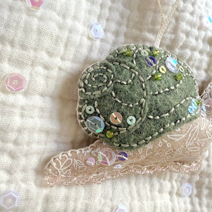 A close-up of a handmade felt ornament shaped like a shell, decorated with white embroidery, iridescent sequins, and small green beads, resting on a cream quilted fabric with lace trim.