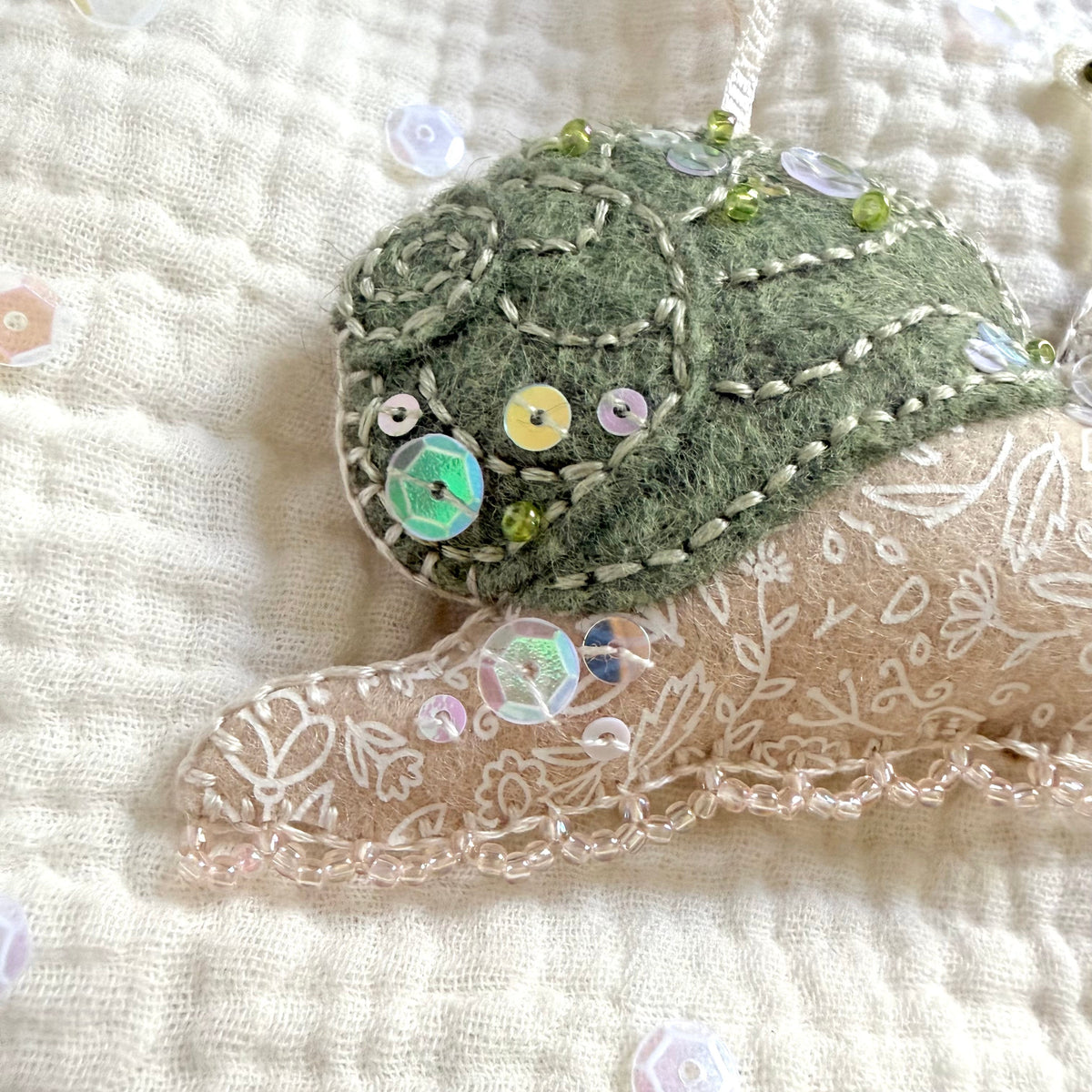 A close-up of a handmade felted wool snail ornament in sage green and beige, embellished with sequins, beads, and green embroidery, resting on a textured cream muslin fabric.