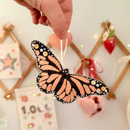 A hand holds a monarch butterfly ornament with orange and black patterned wings decorated with small beads and sequins, against a softly blurred festive background.