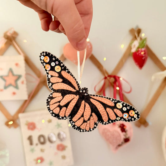 A hand holds a monarch butterfly ornament with orange and black patterned wings decorated with small beads and sequins, against a softly blurred festive background.
