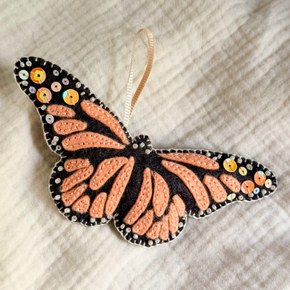 A monarch butterfly ornament with orange and black patterned wings, decorated with small sequins, resting on a textured white fabric background.