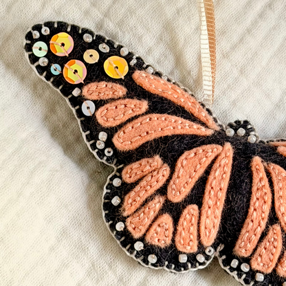 A monarch butterfly ornament made from black felted wool with peach-orange wing patterns, small sequins around the wing edges, and a cream ribbon hanger, shown on a textured off-white fabric background.
